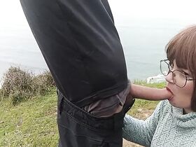 On a cliff overlooking the sea, a brunette beauty sucks her friend's cock